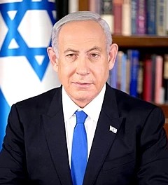 A formal portrait of Israeli Prime Minister Benjamin Netanyahu. He is wearing a dark suit with a white shirt and a bright blue tie. He has a serious expression and is looking directly at the camera. The Israeli flag is visible in the background, along with a bookshelf.